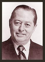 A black and white photo of Senator Joseph Montoya, who is facing the camera and smiling. Senator Montoya wears a white shirt, a black tie and a black jacket.