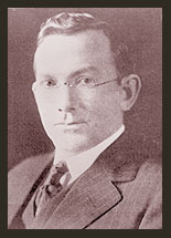 A black and white photo of Senator Sam G. Bratton, who is wearing a grey jacket, a tie, and a white shirt. Senator Bratton wears oval eyeglasses.