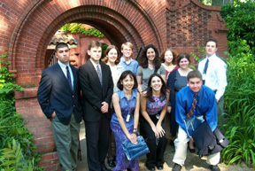 A group photo of eleven young interns, five men and six women, standing before an arched brick doorway and surrounded by green plants.