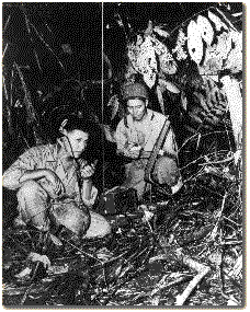 Black and white photo of two young Navajo Code Talkers in a jungle like clearing. One is talking on the radio while the other is taking notes beside him.